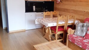 Una cocina compacta con armarios de madera, un refrigerador blanco y una estufa negra. Hay una mesa de comedor con sillas y un sofá rojo con funda estampada con corazones.