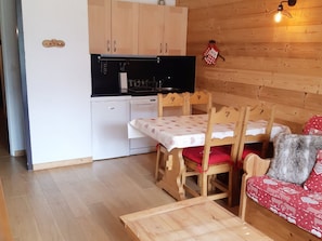 A compact kitchen with wooden cabinets, a white refrigerator, and a black stove. There is a dining table with chairs and a red sofa with a heart-patterned cover. - Studio 4 pers, south balcony, Les Deux Alpes (Les Deux Alpes)