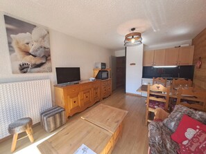 A living room with a television, wooden furniture, a dining area with chairs, and a kitchen in the background.