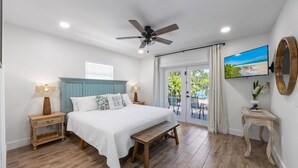 A bedroom with a bed, a ceiling fan, a TV, a wooden bench, and a view of the outdoors.