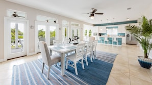 Un grand espace à manger avec une table et des chaises blanches, un tapis bleu et une cuisine en arrière-plan.