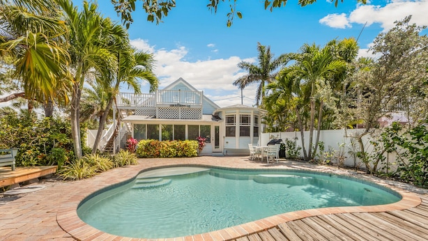 Une piscine dotée d’une terrasse en bois, entourée d’une végétation luxuriante, et une maison avec une véranda en verre.