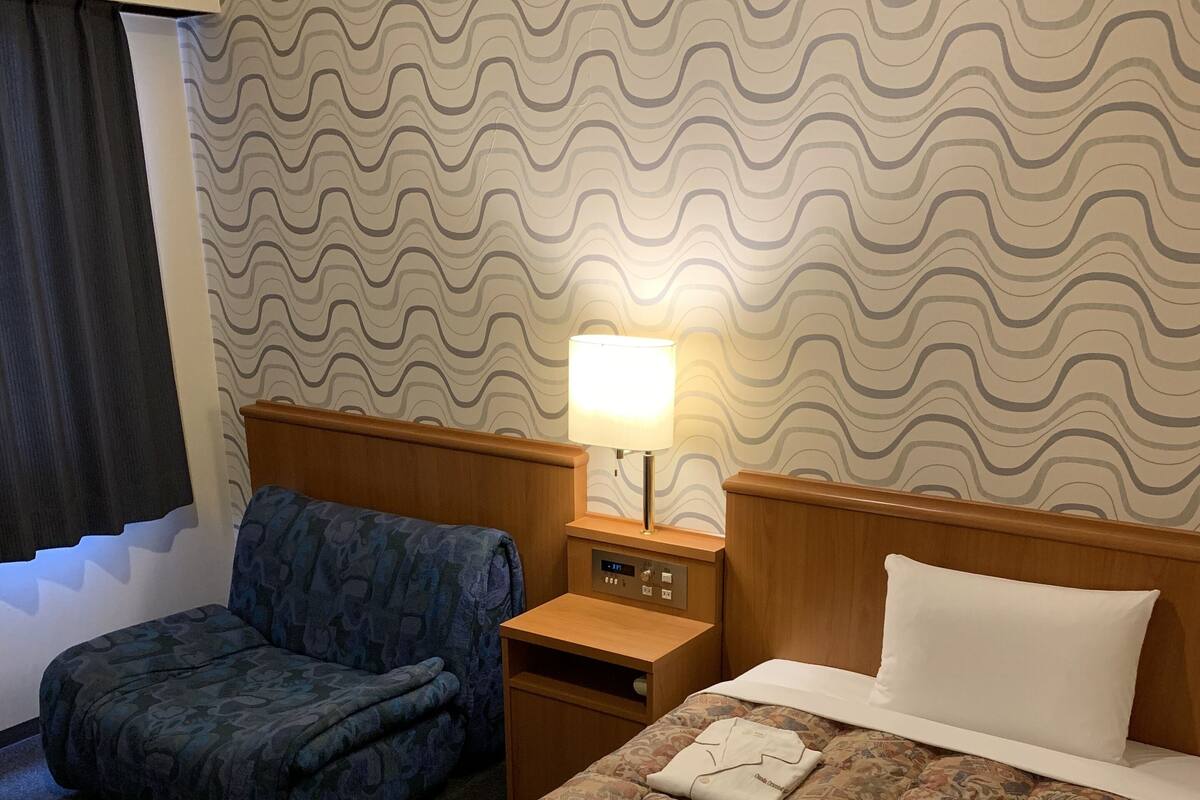 Une chambre d’hôtel avec un lit, une table de chevet, une lampe, une chaise et un mur décoré d’un motif.