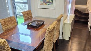 Un coin repas avec une table en bois et des chaises en osier, un tableau encadré au mur et, par une porte vitrée, une vue sur une terrasse.