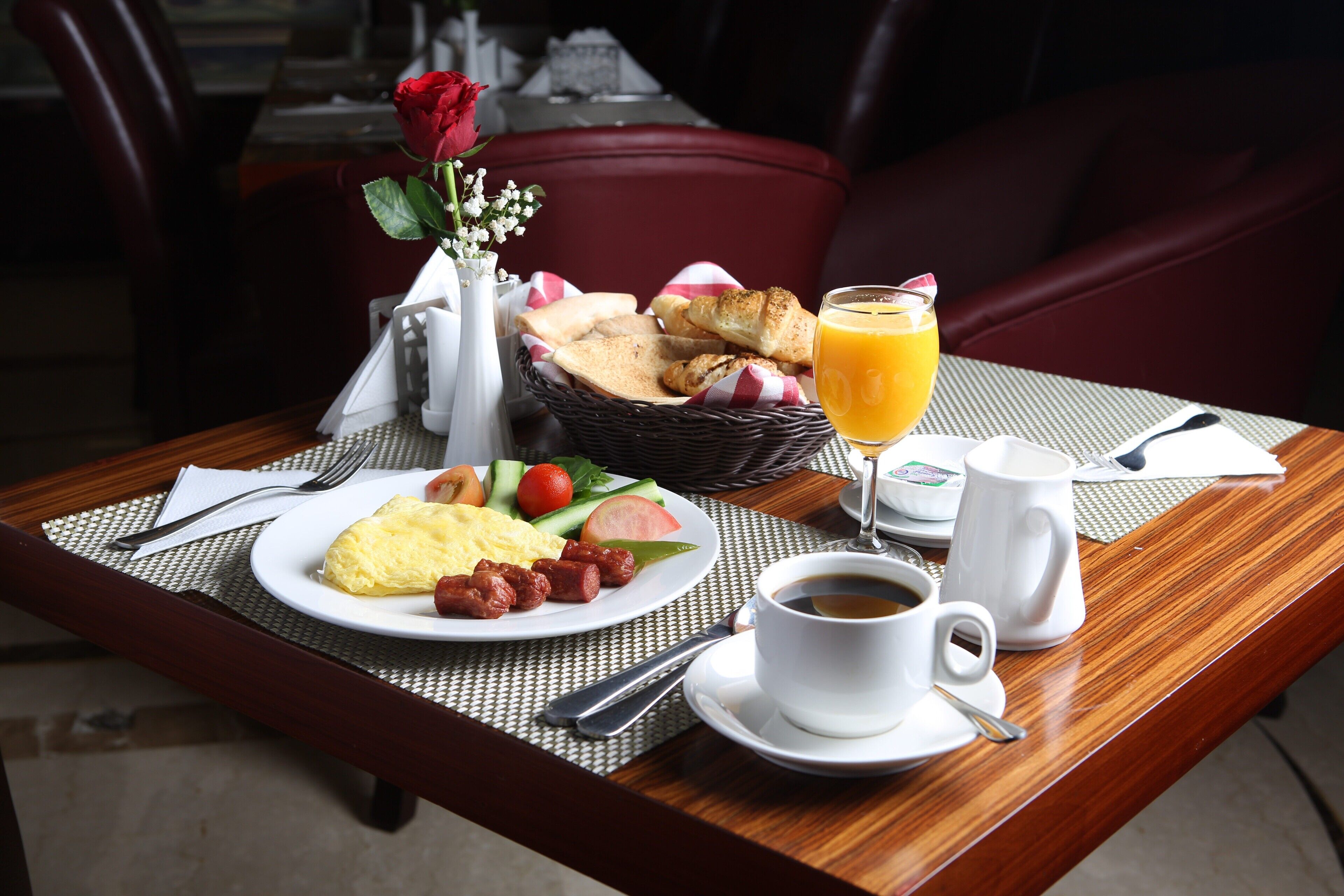 A breakfast table with a plate of eggs, sausages, and vegetables, a glass of orange juice, a cup of coffee, and a basket of bread.