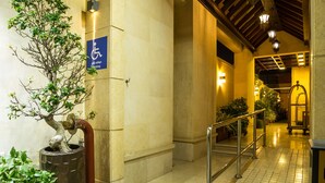 Entrada de hotel con rampa accesible para sillas de ruedas, un árbol en maceta y suelo de baldosas.