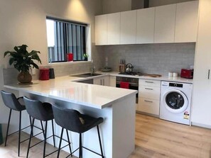 Une cuisine moderne dotée d’un îlot central, de tabourets de bar, d’un lave-linge encastré et d’une fenêtre avec des rideaux.