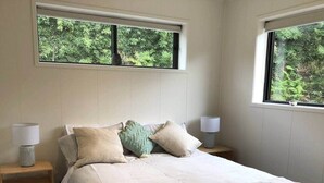 Een slaapkamer met een bed, twee nachtkastjes, twee lampen en ramen met uitzicht op groen.