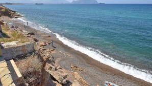 A rocky beach with clear blue water, a few people, and distant mountains.