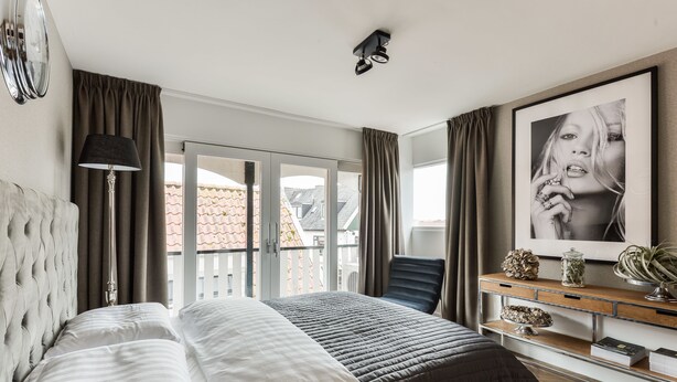 Quarto de hotel com cama, escrivaninha, cadeira, abajur e uma janela grande.
