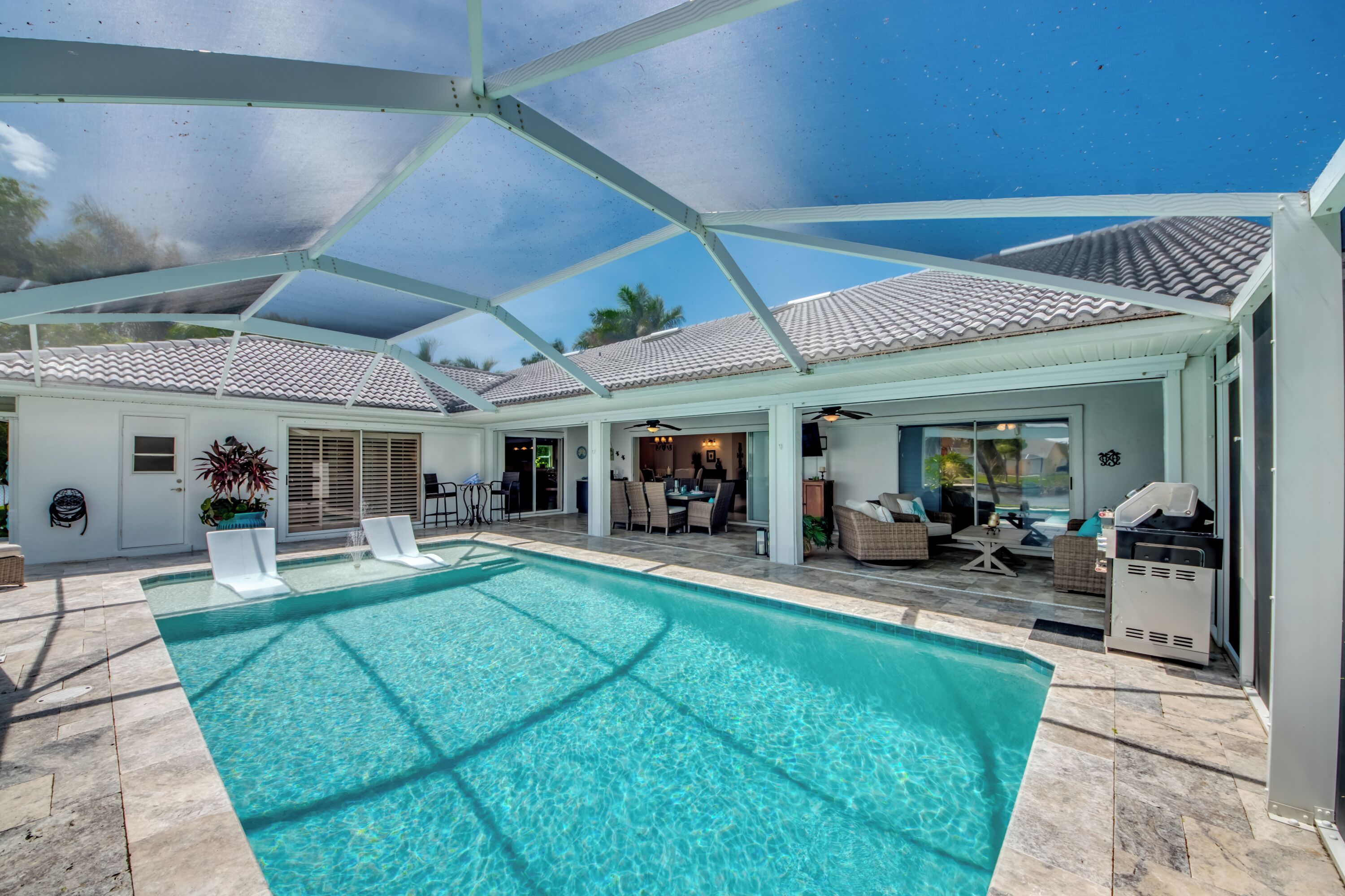 A covered swimming pool area with a patio, lounge chairs, and a grill.