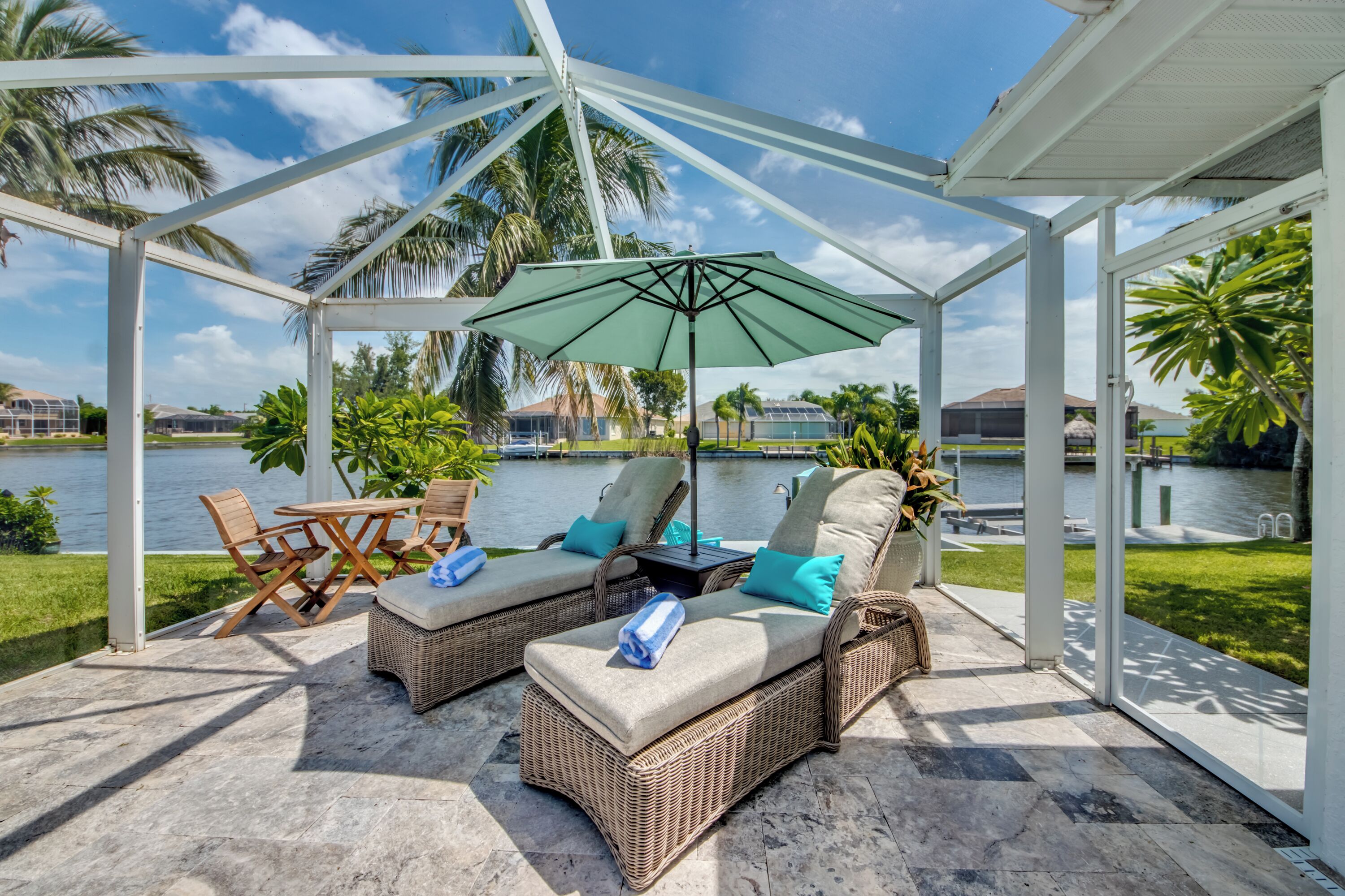 A covered outdoor seating area with wicker furniture, a table, and an umbrella, overlooking a body of water and palm trees.