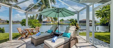 A covered outdoor seating area with wicker furniture, a table, and an umbrella, overlooking a body of water and palm trees.