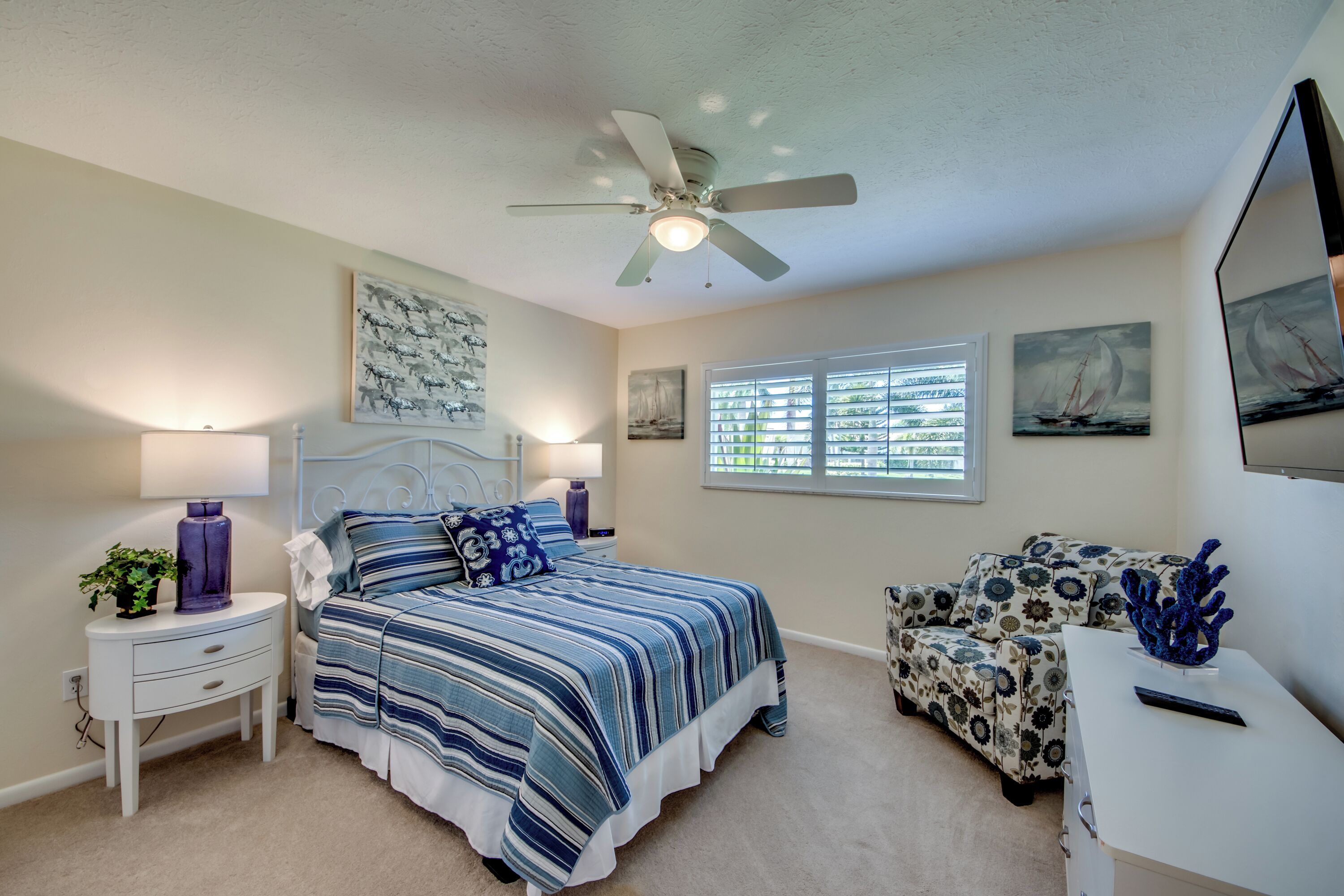 A bedroom with a bed, nightstands, a desk, a chair, a ceiling fan, and artwork on the walls.