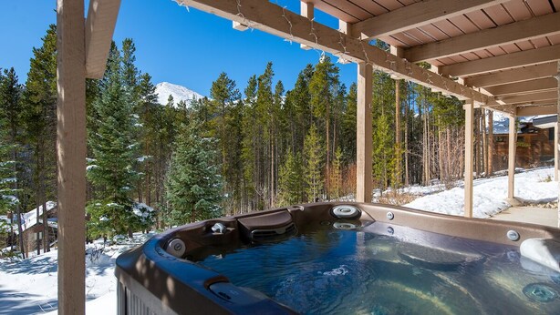 Ett bubbelbad under en träpergola, med en snötäckt skog och berg i bakgrunden.