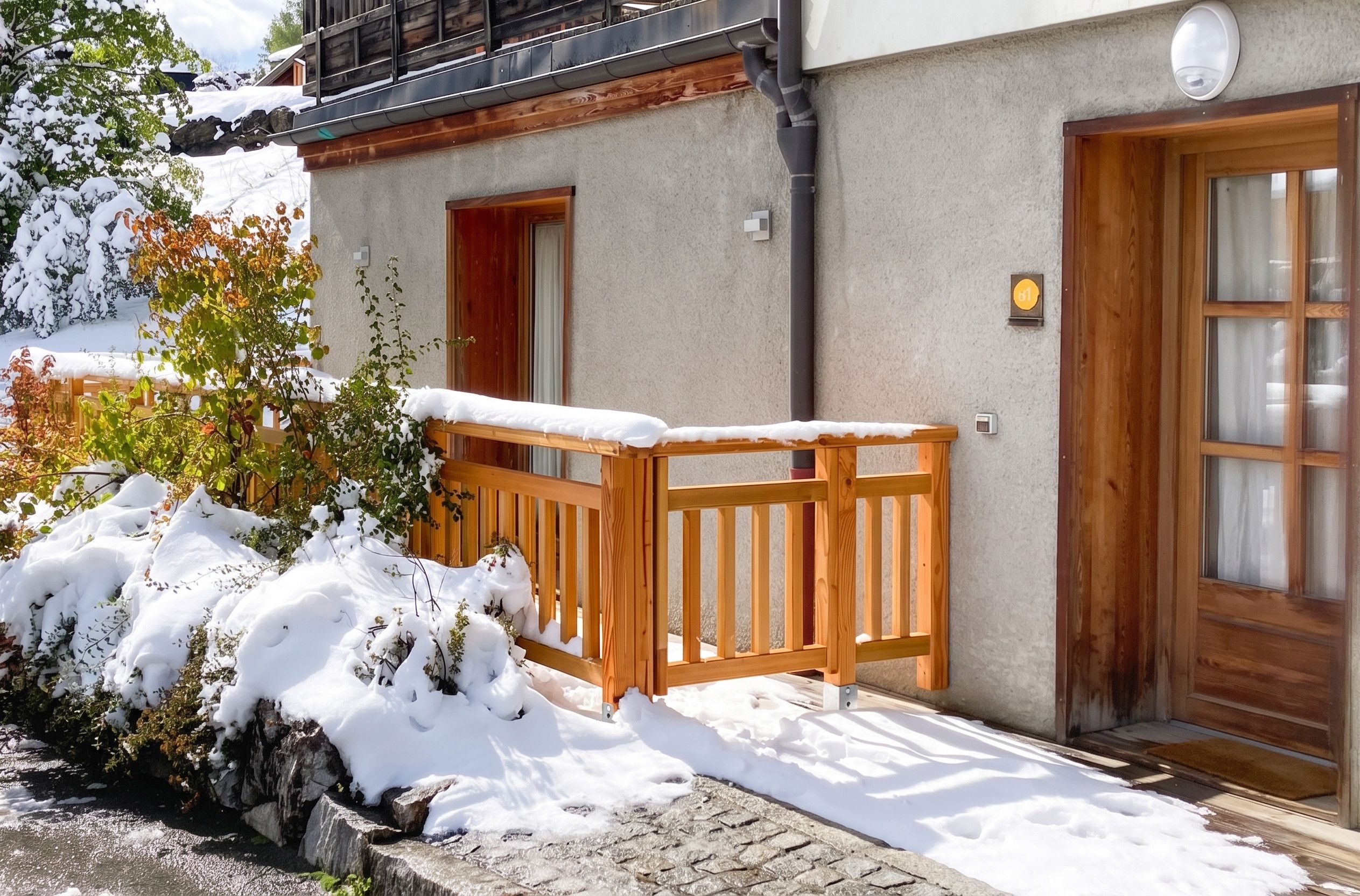 A wooden balcony with a snow-covered ground, a stone wall, and a wooden door with a window.