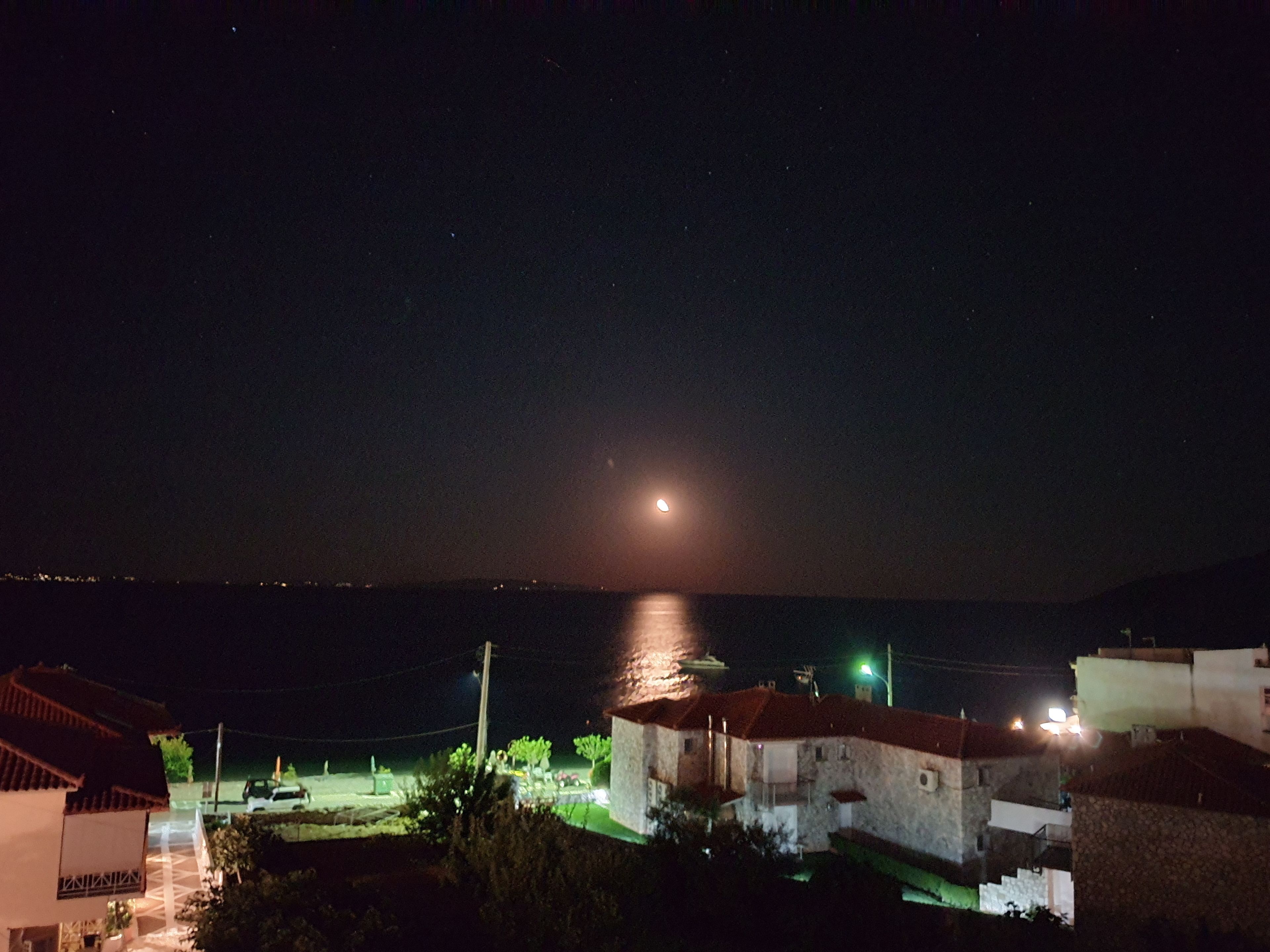 Eine Küstenstadt bei Nacht mit sichtbarem Mond, Häuser mit roten Dächern und einer Wasserfläche.