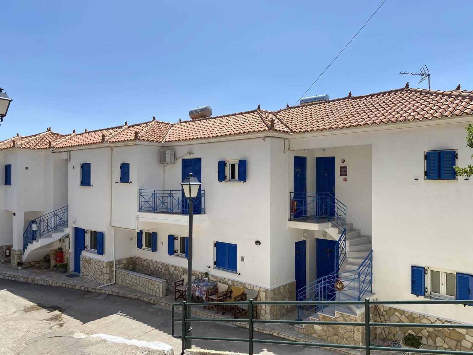 Un edificio blanco con contraventanas azules y techo de tejas. Hay escaleras que conducen a un balcón y una pequeña zona de estar al aire libre.