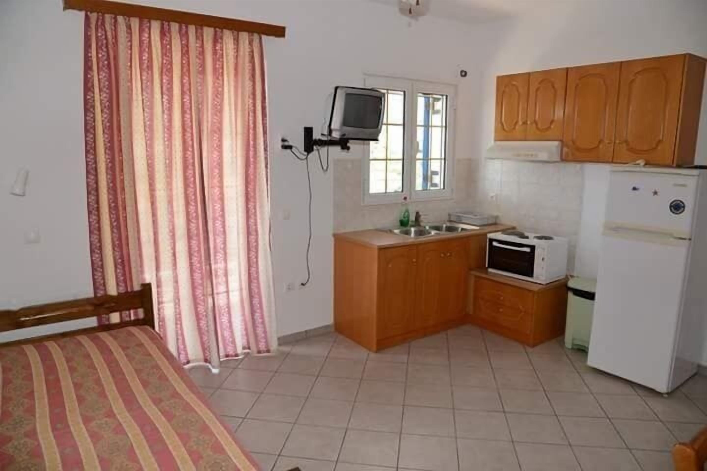 A compact room with a bed, a small kitchenette, a television mounted on the wall, and a window with curtains.