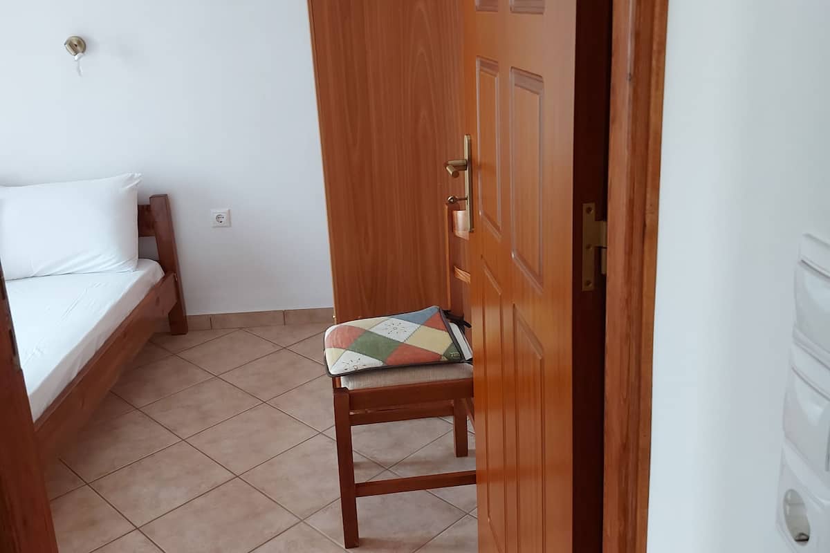 Una habitación con puerta de madera, cama con colchón blanco y una silla con cojín colorido.