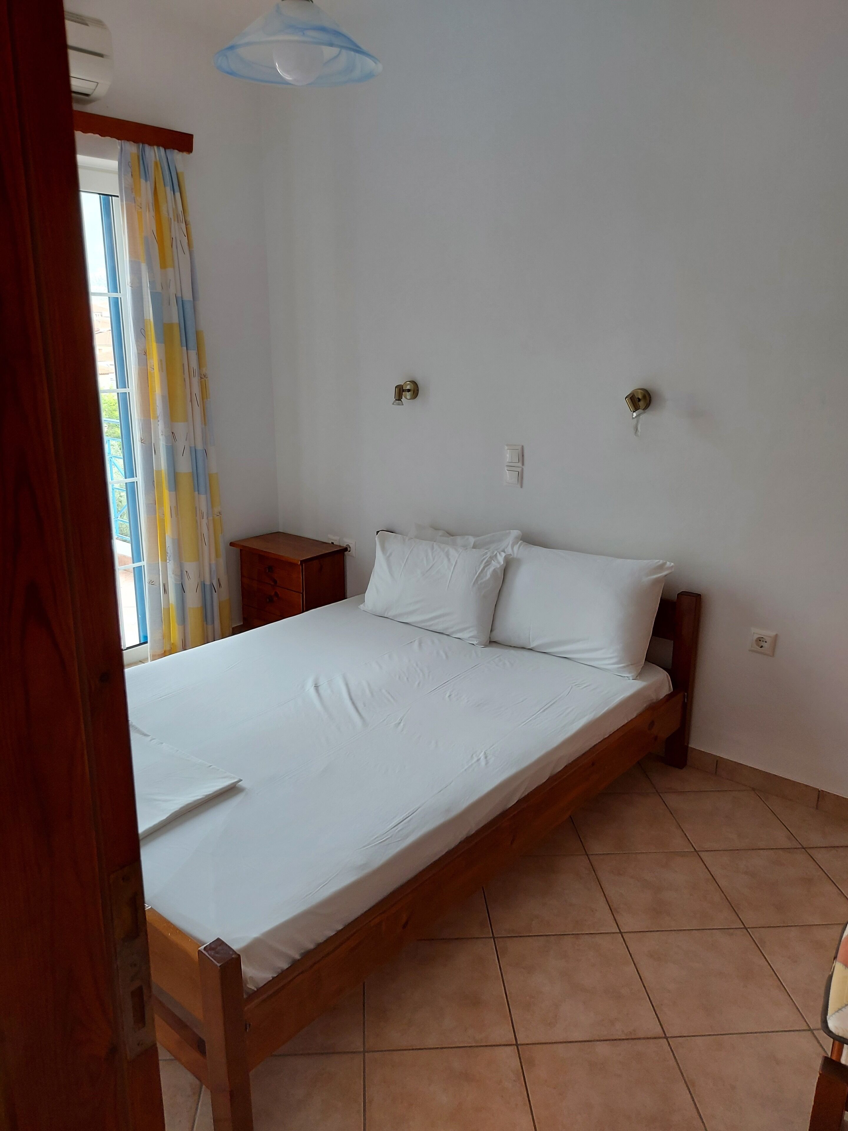 A single-bed room with a wooden frame, white bedding, a small wooden nightstand, a wall-mounted light, and a window with curtains.