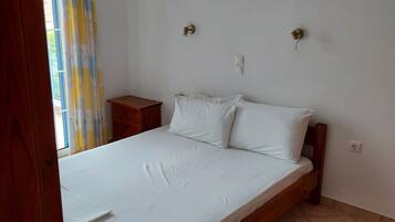 A single-bed room with a wooden frame, white bedding, a small wooden nightstand, a wall-mounted light, and a window with curtains.