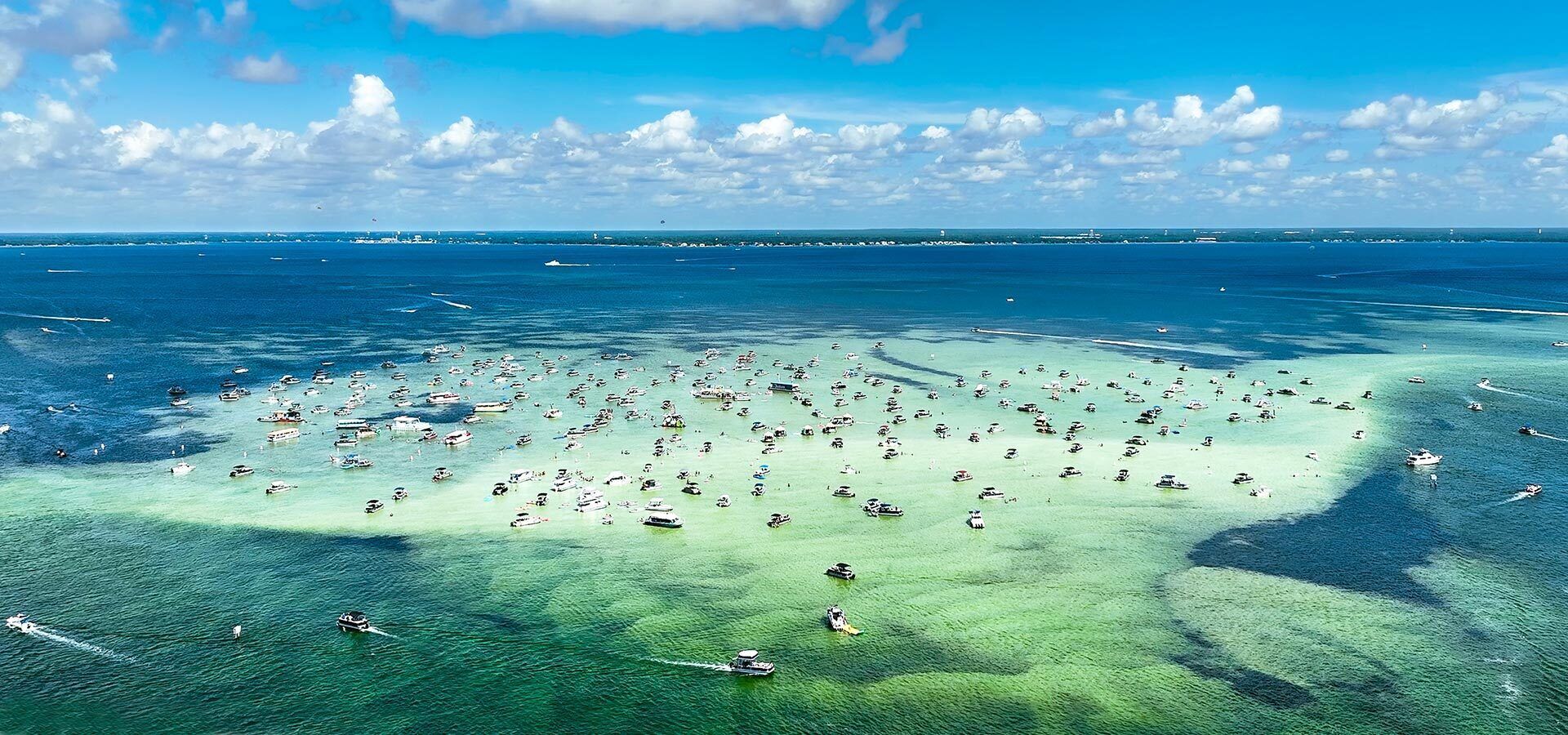 An aerial view of a coastal area with numerous boats and yachts anchored in shallow waters, indicating a popular tourist or recreational spot.
