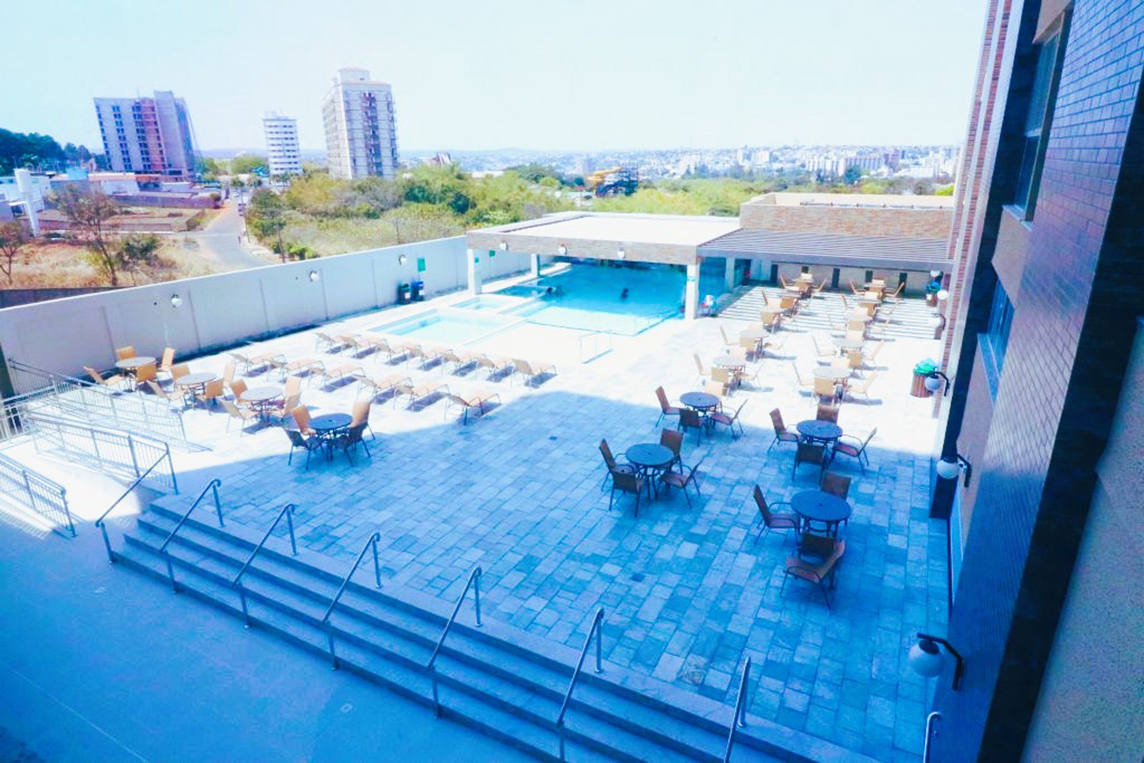 Un área de piscina en la azotea con múltiples sillas de descanso y mesas, una piscina y vistas de edificios de la ciudad al fondo.