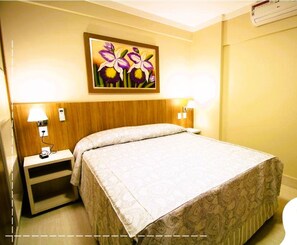 A hotel room with a bed, bedside lamps, a nightstand, a painting of flowers, and an air conditioning unit.