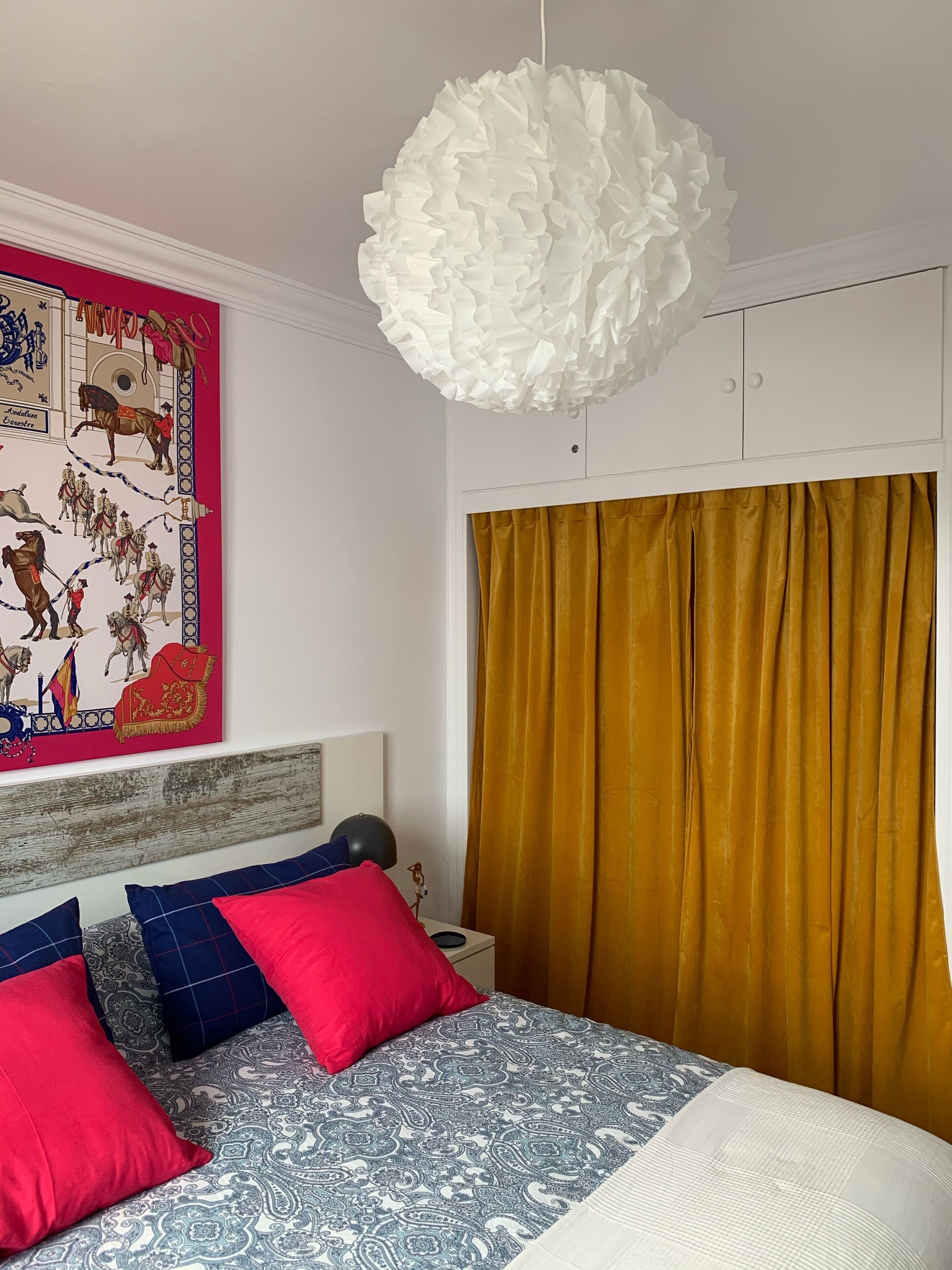A bedroom with a bed, a large ruffled light fixture, a wall with a colorful horse-themed artwork, and mustard curtains.