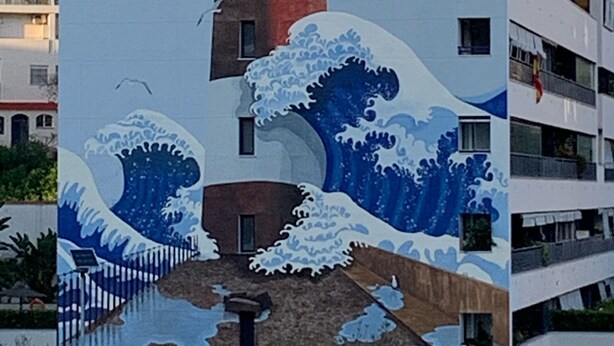 Un immeuble à plusieurs étages dont la façade est ornée d’une peinture représentant des vagues, une tour rouge et blanche, et des balcons.