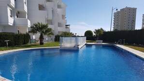 Une piscine à l’eau d’un bleu limpide, entourée d’une terrasse dallée. On aperçoit en arrière-plan des bâtiments blancs, dont l’un possède un balcon et l’autre une façade plate.