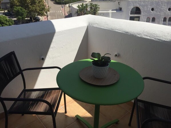 A balcony with a green table and two chairs, a potted plant, and a view of buildings. - Apartment sea view, bright, cozy, facing the port, swimming pool, parking, wifi. (Estepona)