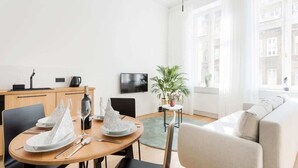 A modern living room with a dining area, a flat-screen TV, a potted plant, and a view of buildings through large windows. - Comfortable Studio in the Center | Close to the Park and the Fair (Poznan)