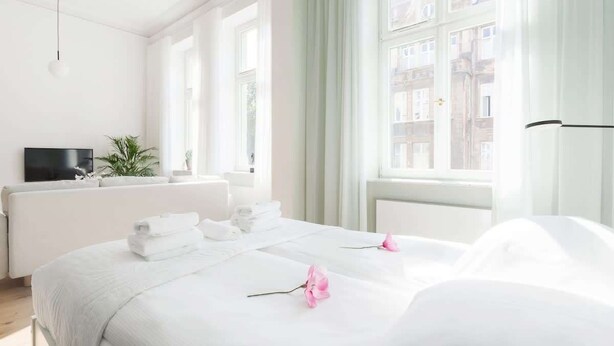 A hotel room with a large bed, a TV, a plant, and a view of buildings through the windows. - Comfortable Studio in the Center | Close to the Park and the Fair (Poznan)