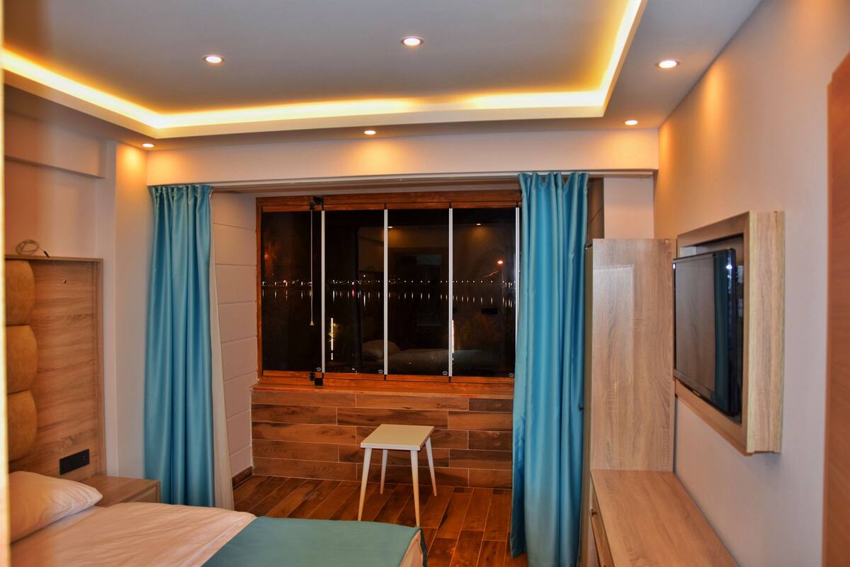 Un dormitorio moderno con un ventanal grande, un taburete pequeño y un televisor en la pared.