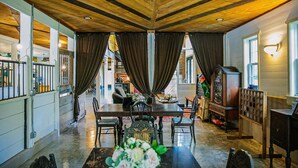 Uma sala de jantar ampla com teto alto e vigas de madeira, com uma mesa comprida com cadeiras, um lustre e uma grande janela com cortinas.