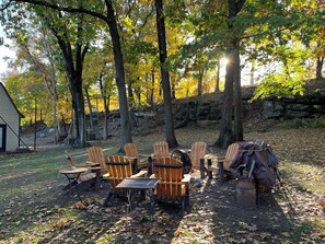 A rustic outdoor seating area with Adirondack chairs and a table, surrounded by trees and a stone wall.