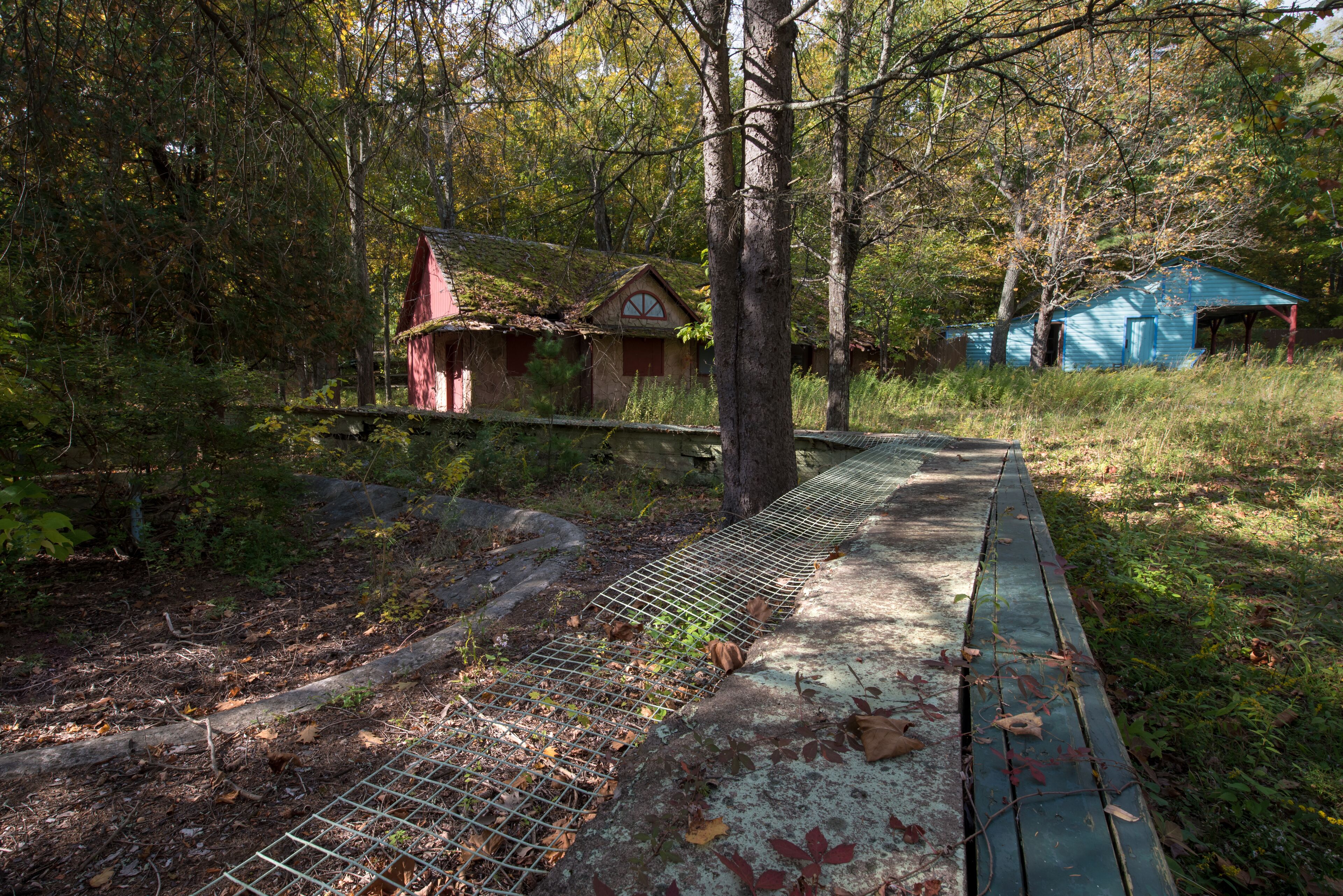 A rustic wooden bridge over a concrete path, surrounded by dense forest and two small cabins.
