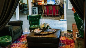 Um quarto aconchegante com uma poltrona verde, um tapete colorido e uma mesa de centro de madeira. Uma escadaria visível ao fundo leva a um espaço de bar.