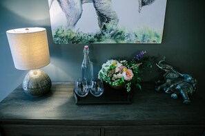 A wooden side table with a lamp, a bottle, two glasses, a floral arrangement, and a small sculpture.