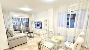 Un salón moderno con una mesa de comedor de vidrio, sofás blancos y un televisor montado en la pared.