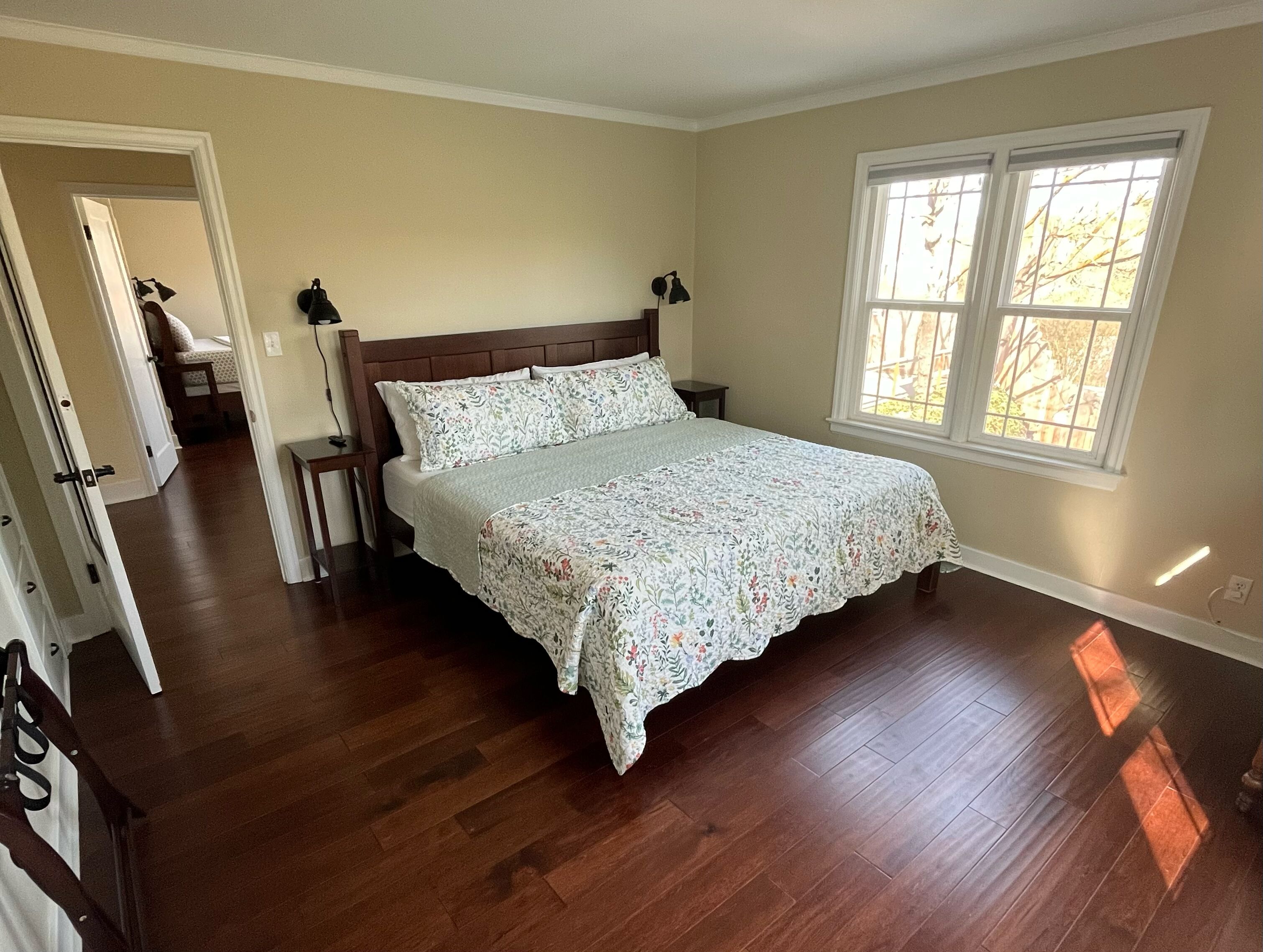 Un dormitorio con cama de madera, una mesita de noche, un buró, una ventana con vistas y una puerta que da a otra habitación.
