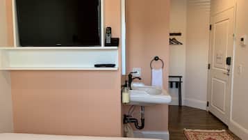 A hotel room with a TV mounted on the wall, a sink with a towel ring, and a small table with a mirror.