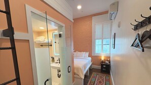 A bedroom with a bed, a nightstand, a lamp, a window with blinds, a coat rack, and a bathroom with a glass door. - The Cabot Lodge (Beverly)