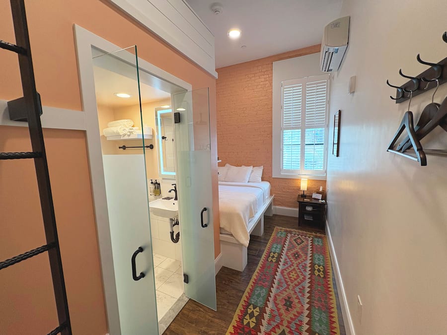 Quarto com cama, mesa de cabeceira, luminária, janela com persianas, cabideiro e banheiro com porta de vidro.