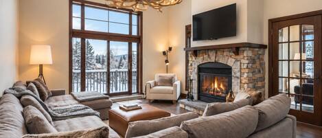 A spacious living room with a stone fireplace, a large window with a view of snow-covered trees, and a modern chandelier.
