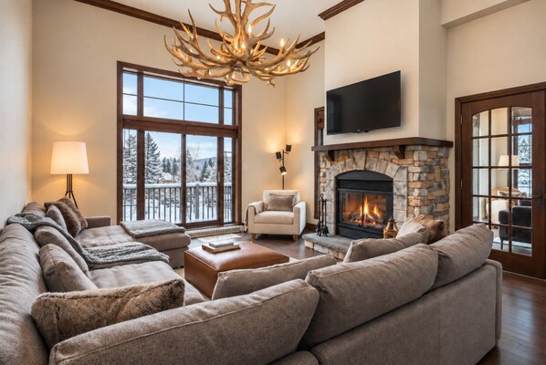 A spacious living room with a stone fireplace, a large window with a view of snow-covered trees, and a modern chandelier.