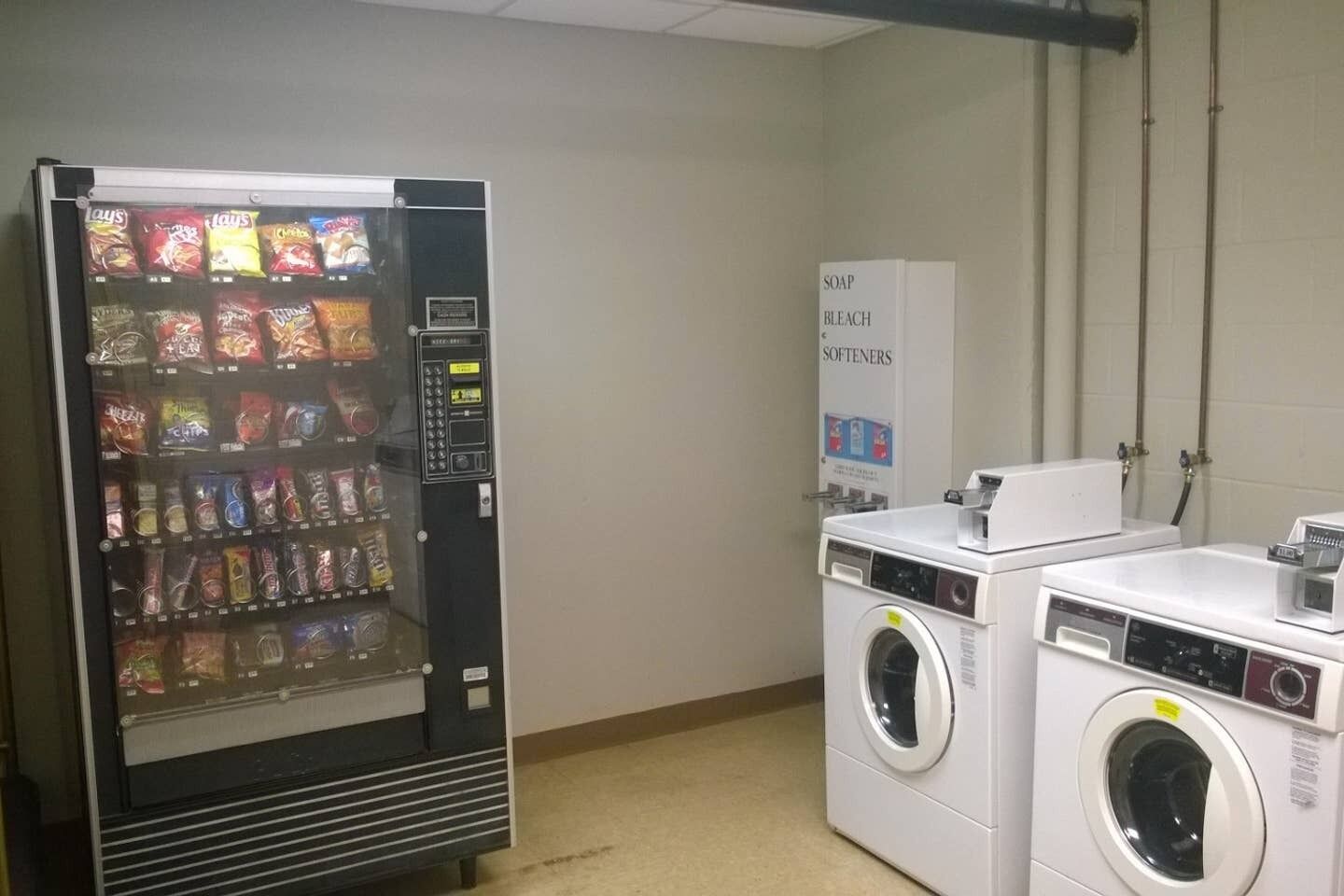 A vending machine with snacks and a washing machine in a room with plain walls and a tiled floor.