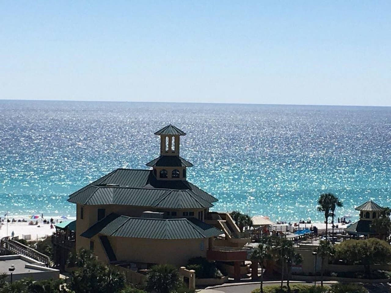Un edificio costero con tejados escalonados y un campanario, con vista a una playa donde hay gente y sombrillas.